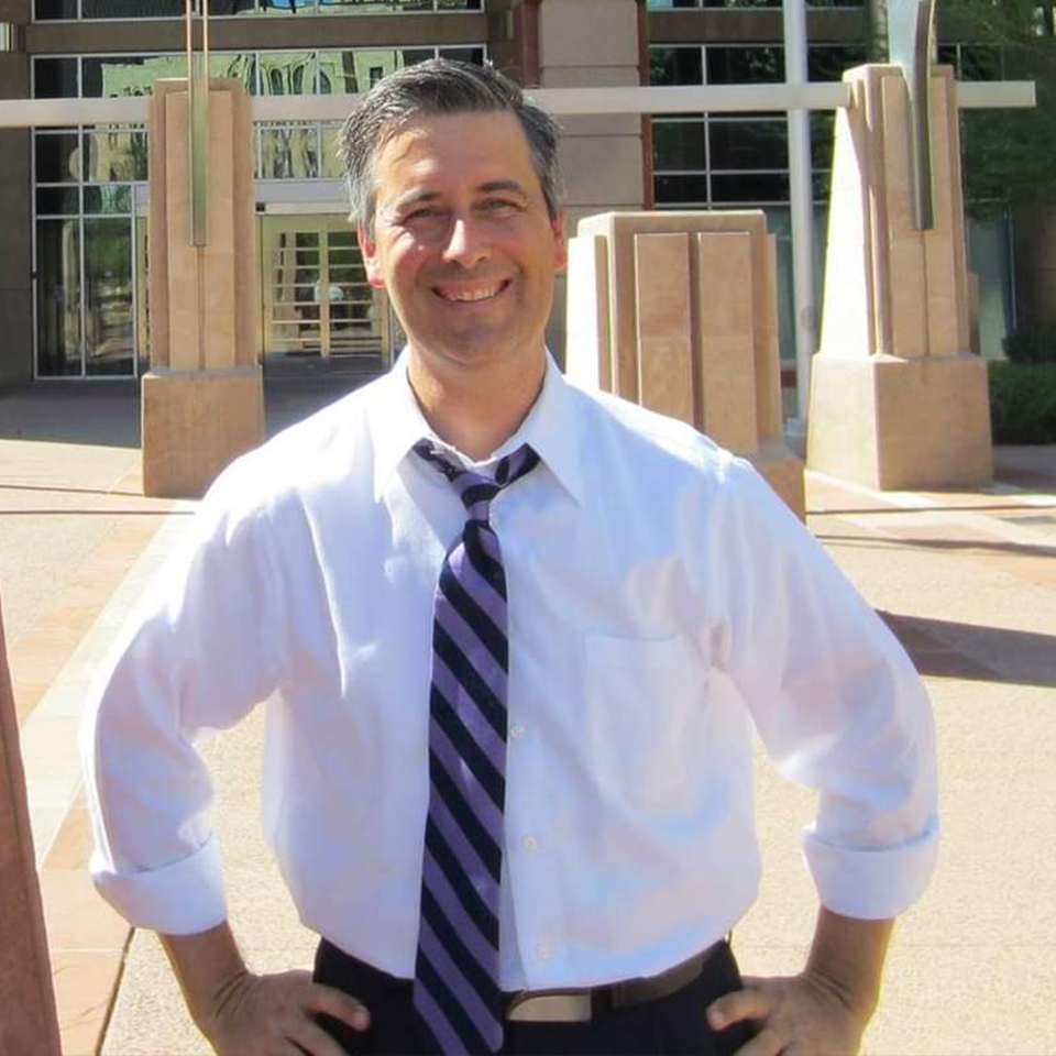 Image of Dan Carroll - Man standing outside in white shirt and tie, hands on hips.